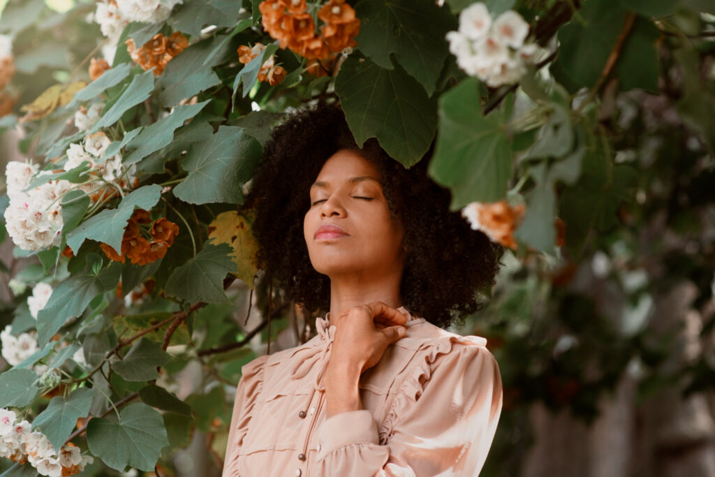 A serene woman in a neutral-toned outfit stands under lush green foliage and flowers, her eyes closed in a moment of calm reflection. The image symbolizes the mental health benefits of making sustainable fashion choices.