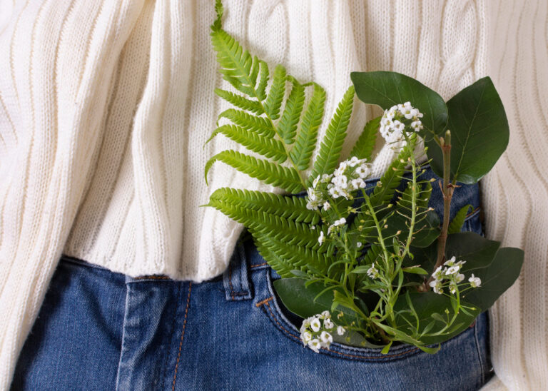 A white knit sweater tucked into blue denim jeans with green leaves and small flowers emerging from the pocket, symbolizing sustainable and eco-friendly fashion.