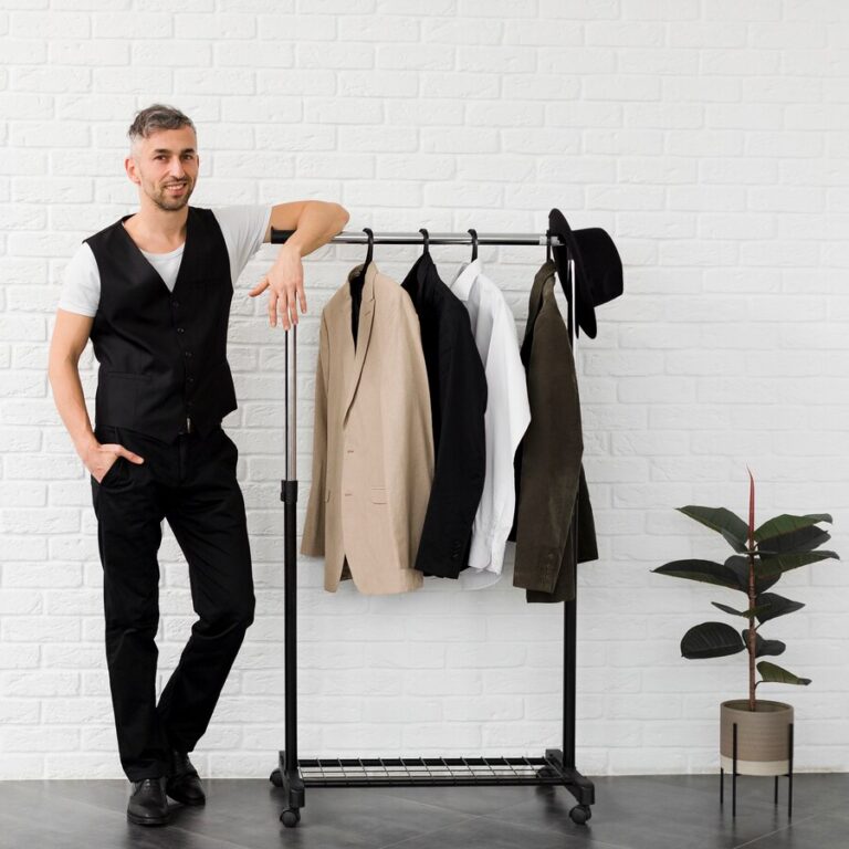 Man standing beside a minimalist wardrobe rack with neutral-toned blazers and shirts, set against a white brick wall with a potted plant.