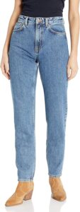 Model wearing Nudie Jeans Women's Breezy Britt high-waisted straight-leg jeans in classic blue, made from organic cotton, paired with a black top and tan heeled ankle boots, standing against a white background.