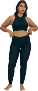 A person with medium-toned skin and dark hair is shown from the chest up, modeling black Pact organic cotton leggings and a matching high-neck crop top. They are barefoot and standing against a plain white background, with hands on the waistband of the leggings.