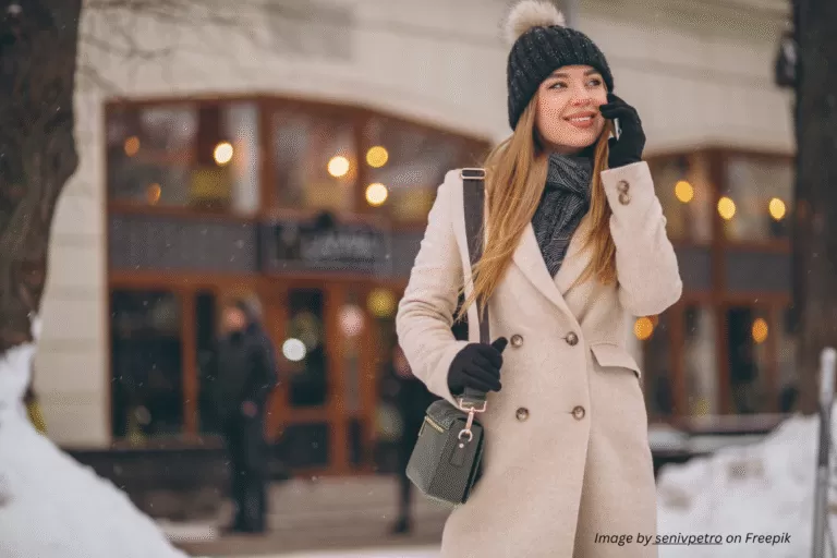 A woman in a cream sustainable wool coat smiles on a snowy city street, styled with a black knit hat and eco-leather crossbody bag — a timeless sustainable coat look for winter.
